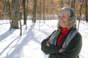 smiling white woman with shoulder-length silver hair, wearing a red turtleneck shirt, dark jacket, and striped grey scarf, outdoors on sunny snowy day