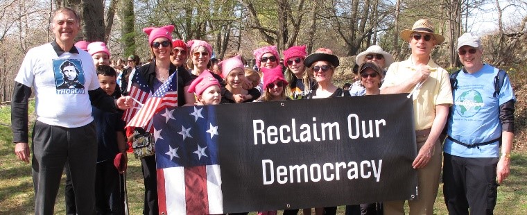 group of people, a couple holding USAmerican flags, current and 13-star, some wearing pink pussy hats from Women’s March 2017, hold a sign reading Reclaim Our Democracy, with a folded USAmerican flag on the left side