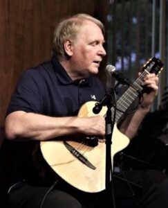white blond middle-aged man in a black short-sleeved shirt playing a guitar at a microphone