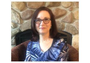 white woman with shoulder-length dark hair, brown glasses, and a blue-and-white patterned shirt sits in a brown upholstered chair in front of a stone fireplace