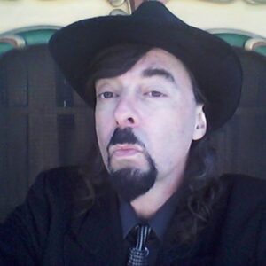 White man dressed in black shirt, tie, and jacket, with long black hair, moustache, and beard, wearing a black fedora hat