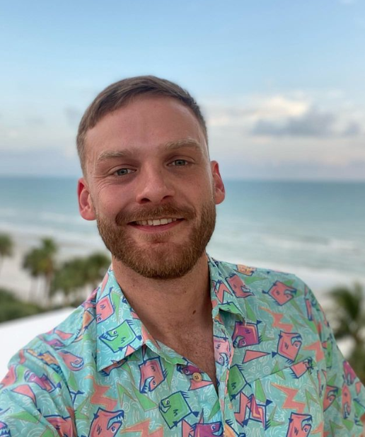 Smiling white adult man with light brown hair and beard wearing a colorful shirt with a beach in the background
