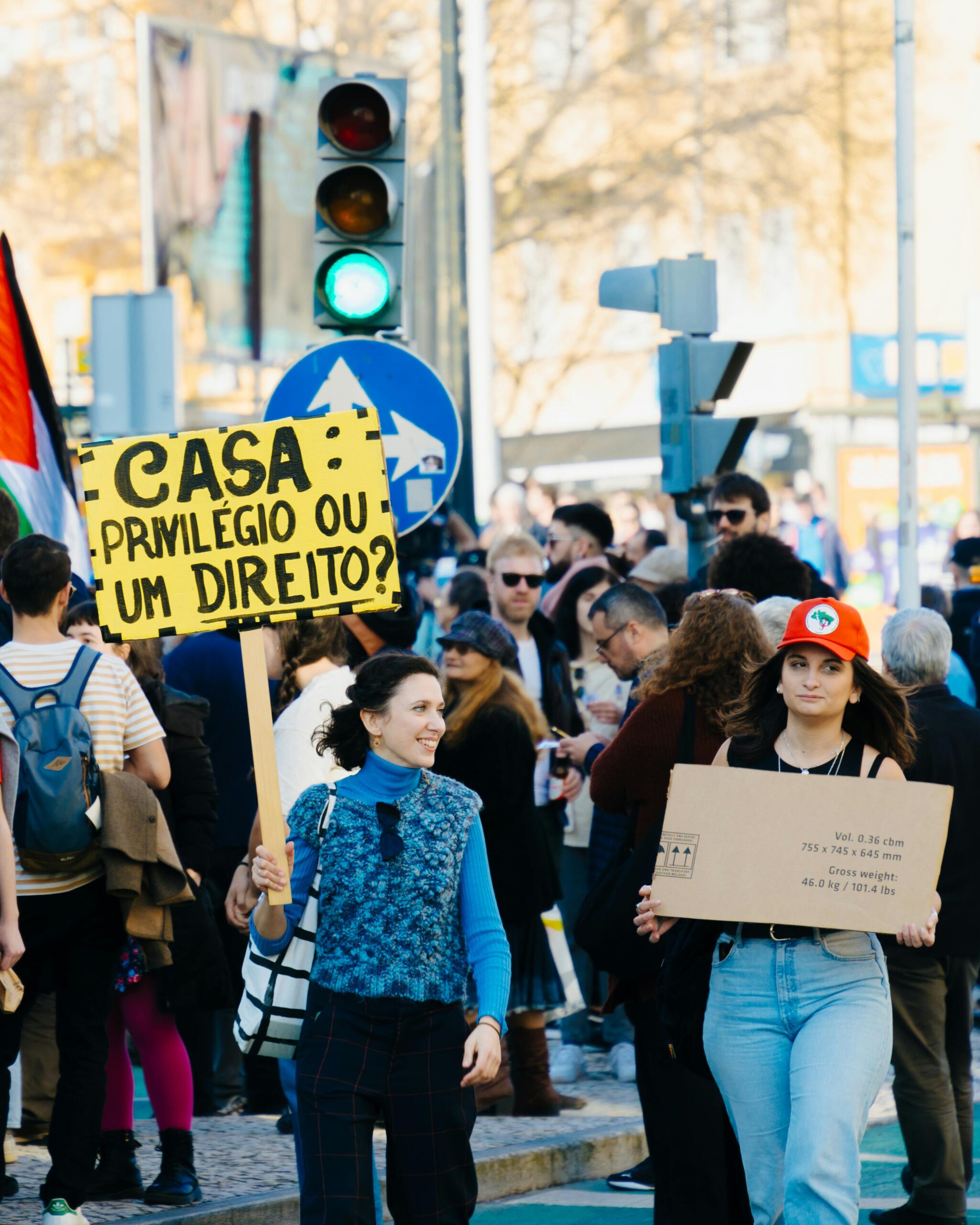 Crowd in front of streetlights, 2 women in front holding signs, one saying Casa: privilégio ou um direito?, the other a piece of a brown cardboard box.