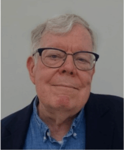 White man with grey hair and dark glasses wearing a blue shirt and dark jacket.