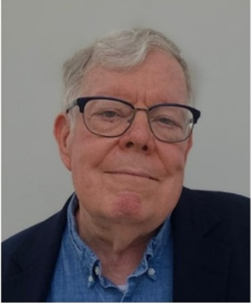 White man with grey hair and dark glasses wearing a blue shirt and dark jacket.
