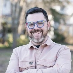 smiling young white man with salt-and-pepper beard and moustache wearing blue glasses in an outdoor setting