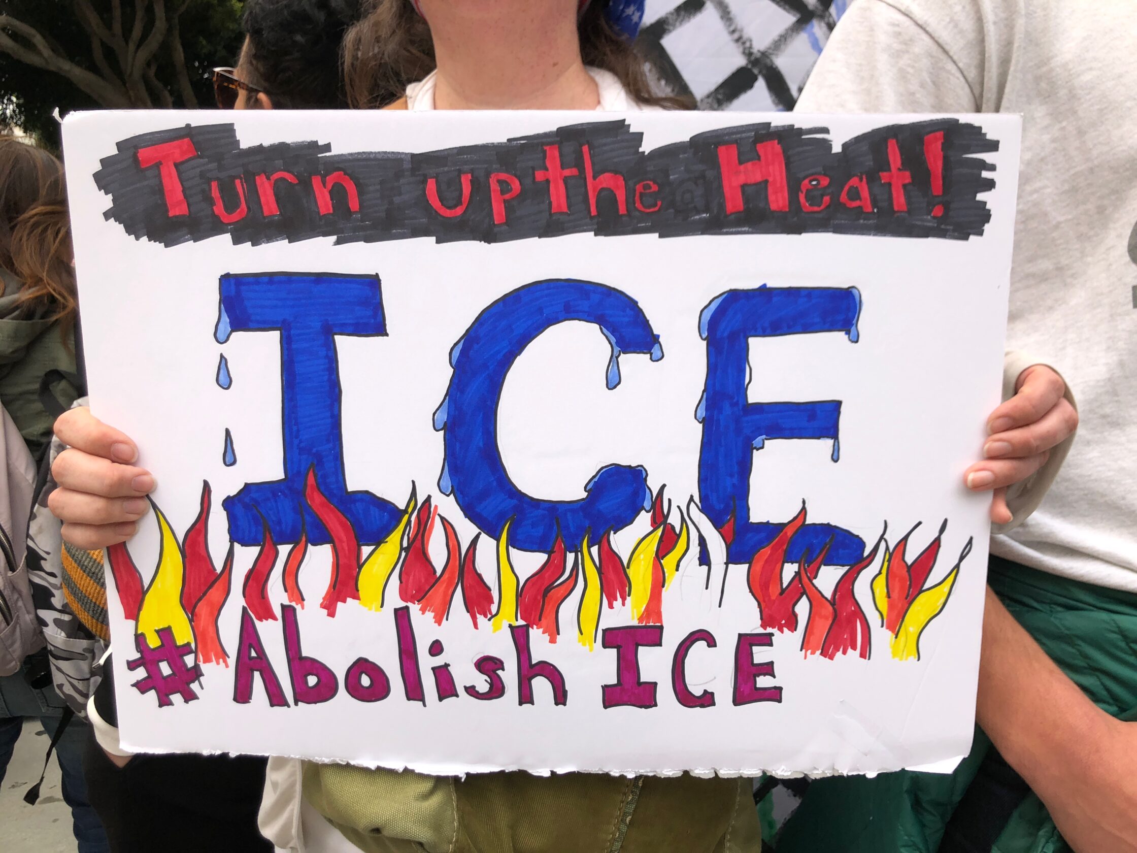 Colorful poster held up at protest reads at top, “Turn up the Heat!”, at bottom, “#Abolish ICE” Between these lines, “ICE” in blue, with water drips starting to fall off, is above red, orange, and yellow flames.