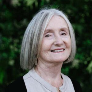 smiling white woman with grey hair, shoulder-length straight hair and dark eyes wearing a white collarless shirt and dark jacket